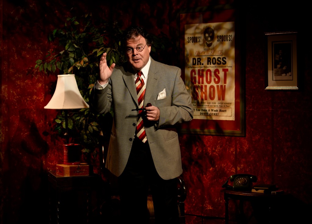 John Carney in character, wearing glasses and a striped tie, on a theatrical set